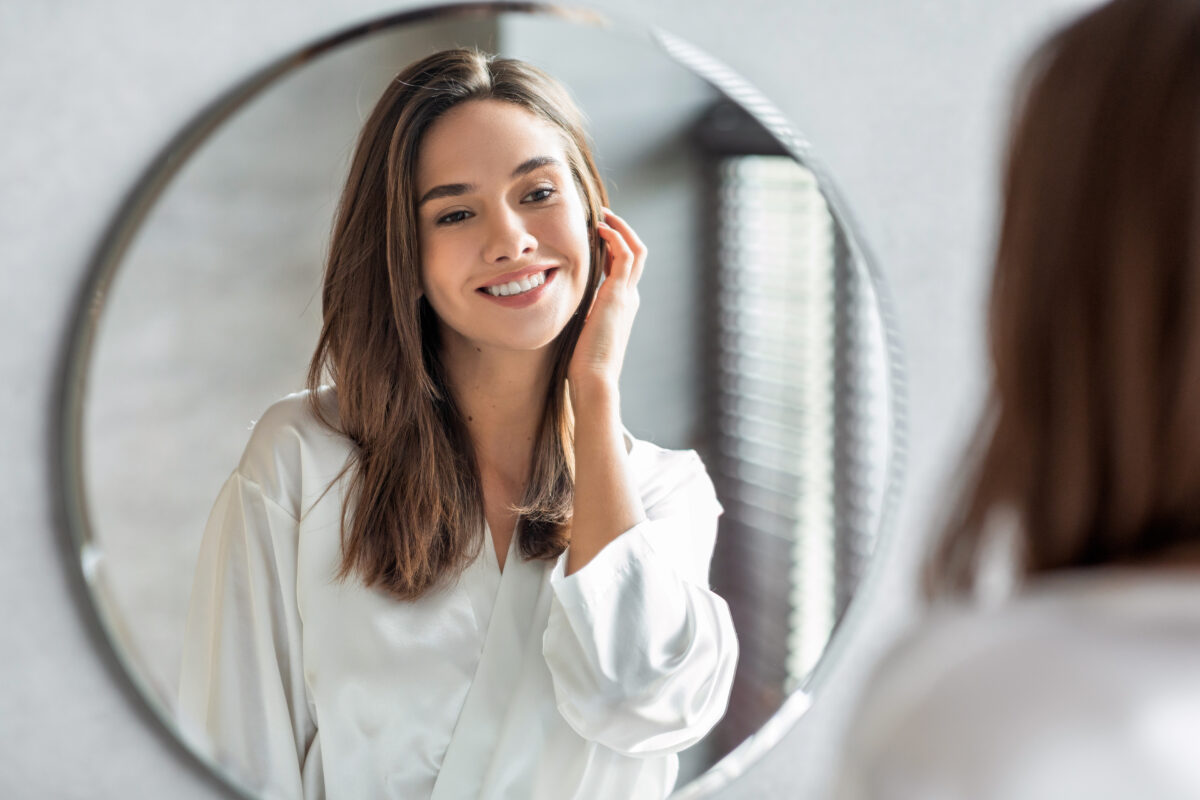 Skönhetskonceptet. Porträtt av attraktiva lycklig kvinna tittar på spegeln i badrummet, vacker Millennial Lady bär vit silkeskåpa leende till reflektion, njuta av hennes utseende, selektivt fokus